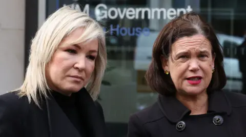 PA Media Sinn Féin deputy leader Michelle O'Neill (left) and leader Mary Lou McDonald (right). O'Neill has blonde shoulder-length hair and McDonald has a dark brown bob. Both are wearing black coats.