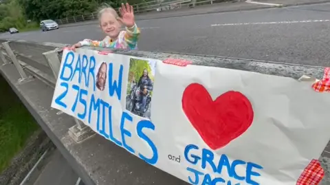 Girl waves to bikers with sign that says '275 miles to Barrow'