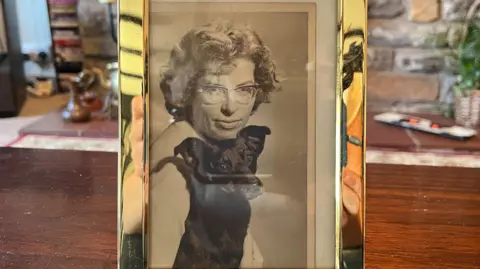 A black-and-white photograph of Iris Sibley in a gold photo frame. She has curly hair and is wearing glasses as she looks at the camera. She is sitting with a black dog.
