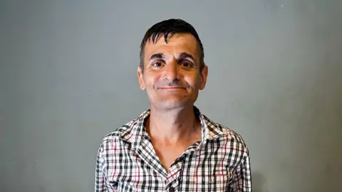 DAVE EDWARDS/BBC A 47-year-old man looks into the camera with a smile. He has dark hair and is wearing a checked shirt.
