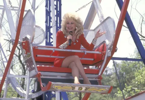 Ron Galella, Ltd./Ron Galella Collection via Getty Images Dolly Parton wearing a bright red jacket and skirt with leopard print cuffs, sitting on a ferris wheel, smiling and speaking into a microphone