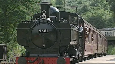 BBC Archive Picture of a black steam train carrying multiple carriages. The train has the conductor and passengers leaning out the window as it approaches the station. The train is surrounded by trees and bushes. 