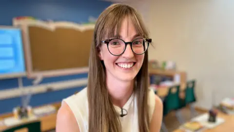 Jess Mancini is smiling at the camera. She is wearing glasses and has long brown hair and is wearing a white sleeveless top.