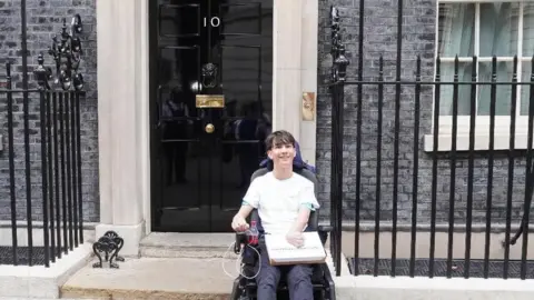 Lucas Lucas is sitting in his wheelchair outside the black front door of 10 Downing Street. He is wearing a white T-shirt and jeans and is smiling at the camera.