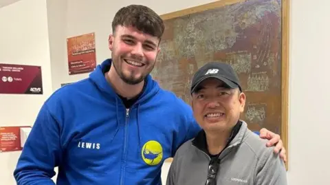 A tall young man in a blue hoodie holds two boxes of chocolates in one hand with the other outstretched over the shoulder of a smaller man wearing a black cap and grey hoodie. They are standing in a gym communal area while smiling for a photograph.