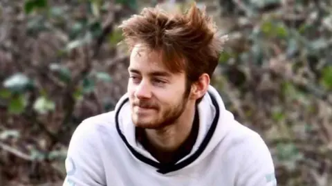 Toby Parker, 24-year-old motorcyclist - he is sat down outside wearing a white hoodie with black piping - he is looking to the left hand side and has short brown hair and a beard.