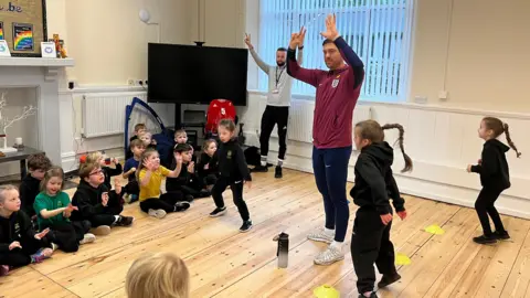 Jack Rutter and a PE teacher stand up in a primary school hall with their hands in the air as about 12 children sit down and watch others do star jumps, with yellow plastic cones marking the spot they are to stand in on the light coloured wooden floor