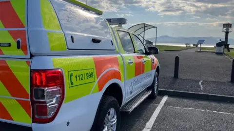A white, orange and yellow emergency services van, parked at a carpark, with water and mountains on the horizon
