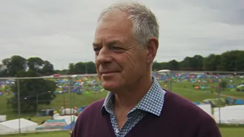 Mr Lane Fox is wearing a burgundy jumper over a chequered blue shirt. He has grey hair, a receding hairline and appears tanned. He has wrinkles on his forehead and around his eyes. In the background one can see festival-goers setting up tents on the grounds.