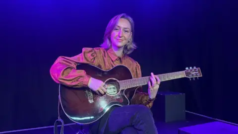 A woman with blonde, shoulder length hair, wearing an orange silky blouse and blue denim jeans. She is on a stage, sat cross legged, holding a brown guitar and smiling toward the camera.