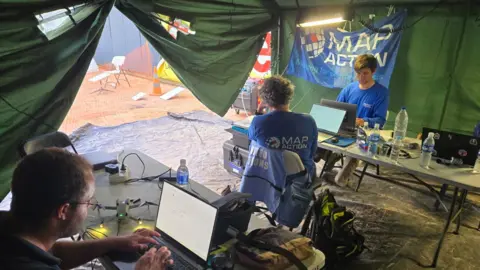 MapAction MapAction workers using laptops inside a field tent, with equipment, water bottles and a MapAction banner visible in the background.