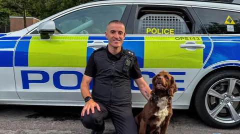 A picture of a police officer kneeling down infront of a police car with a brown spaniel police dog
