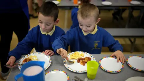 Boys wearing school uniform eat a plate of eggs and sausages in a school canteen.