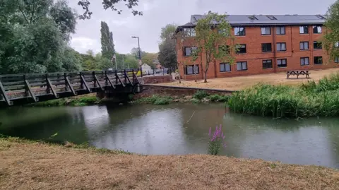 University of Buckingham A river with a wooden footbridge over it and a red brick building in the background. There is a road in the background with trees nearby.