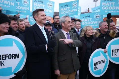 PA Media Matt Goodwin and Nigel Farage stand in front of a crowd, some of whom are holding Reform party placards. Farage is pointing at Goodwin 
