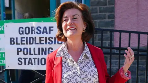 PA Media Eluned Morgan stood in front of a sign that says polling station in English and Welsh. She is wearing a red jacket, a white and black shirt with patterns and is smiling to the camera.