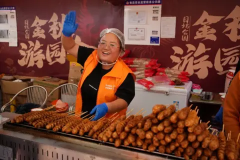 A woman working at a food stall is waving to someone off-camera.