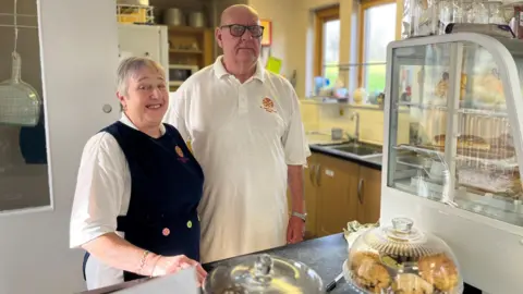 BBC Anne Kemp smiling at the camera next to her husband Derek Kemp behind the counter at Bede's Bakehouse. They are wearing white t-shirts with the red and yellow logo of St Peter's Church. Anne has short grey hair and is wearing a dark blue apron. Derek has black-rimmed glasses. There is a range of cakes and bakes in front of them.