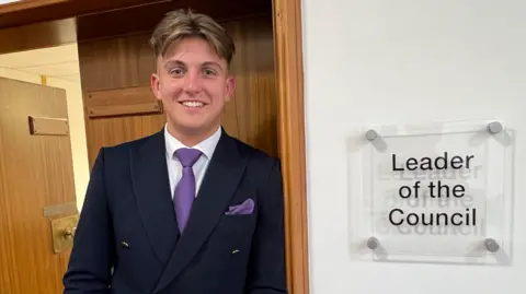 BBC A man in a dark jacket, purple tie and white shirt is next to a door frame on the left of the photo. The sign on the right reads Leader of the Council.
