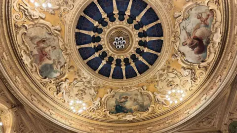 The ornate designs of the ceiling of the Gaiety Theatre, its is mainly coloured cream and yellow. There are drawings featuring mythical beings, such a mermaid and a cherub.