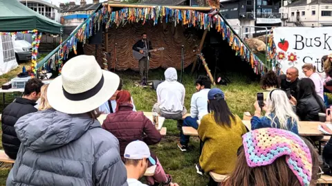 People are watching the Buskers Showcase Stage at a previous Strawberry Fair event. The stage is a half moon type tent - open at the front and closed at the back to give the musician cover. It is on the ground and the crowds of people watching are sat on wooden picnic tables.