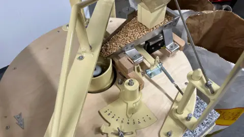 John Devine/BBC A milling machine, wheat grain can be seen going down a triangular chute in where the millstone is, there is a wooden circular cover on the machine with various light yellow metal parts, arms, springs and a numbered gauge.