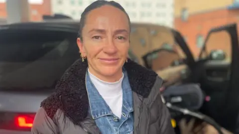 A woman smiles into the camera. She has dark hair tied back and is wearing gold earrings, a black coat with a black fluffy hood, blue denim shirt and white turtleneck. There is a vehicle behind her with the fuel pump connected to the car and driver side door open.