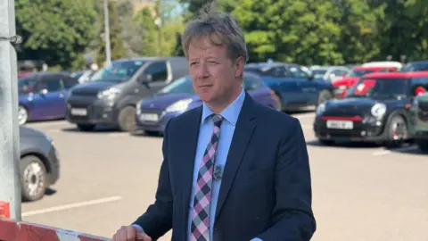 Cambridgeshire and Peterborough Combined Authority Paul Bristow wears a navy suit, light blue shirt and chequered tie. There are parked cars in the background. 