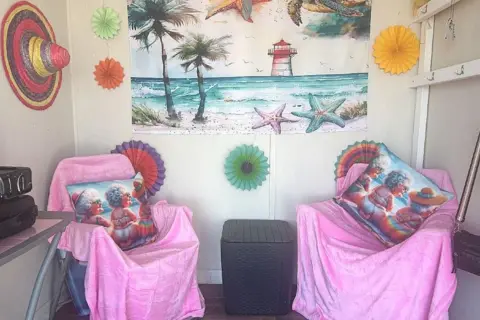 The interior of Liz Symons' beach hut. There are two beach chairs with arms both covered with light bright pink velvet. Both have a cushion with a printed picture of three cartoon drawings of three older women with grey hair wearing bikinis and pictured sitting down from behind on a beach. On the back wall are six brightly coloured fan decorations placed around a printed material drawing of a beach with palm trees, a lighthouse and oversized starfish. On the left wall a Mexican hat in bands of pink, green and natural coloured straw is hanging. The edge of a table can be seen next to it. Between the chairs is a dark brown plastic container which could be used as a side table. There are hooks with keys hanging from one on the right wall.