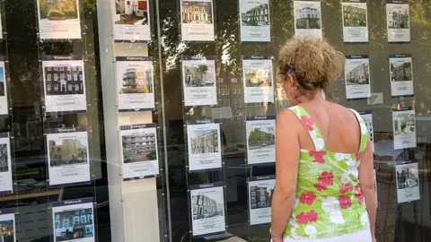 Getty Images Woman in floral sleeveless top looks in estate agent window at rows of listings