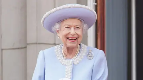 Queen Elizabeth II watches from the balcony of Buckingham Palace during the Trooping the Colour parade on June 2, 2022. She is wearing a lilac outfit with a white rimmed lilac hat. 