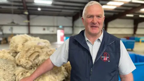 A grey-haired man, wearing a blue jacket and white shirt, stands in front of wool fleeces