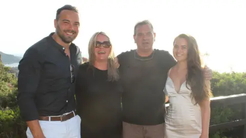 Lucie Macleod with her family looking at the camera smiling. They are standing outside with greenery and the sea behind them. 