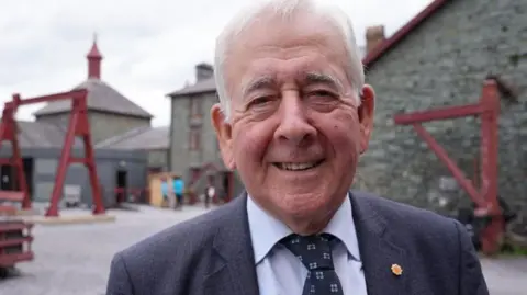 Smiling at the camera, a headshot of a white haired man, wearing a dark suit, blue shirt and navy tie, with a Plaid Cymru badge