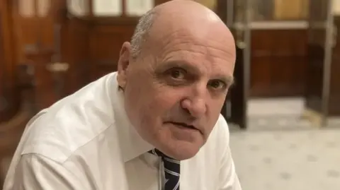 BBC Steve Radford, who is bald and is wearing a white shirt and tie, sits in Liverpool Town Hall 