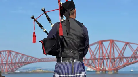 Sacha Previn Kumar A bagpiper, wearing a full kilt playing his instrument in front of the Forth Bridge.