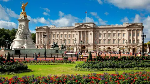 Buckingham Palace on a sunny day.. A three-storey pale grey building in the neo-classical style. In the foreground is a garden with lawns and flower beds. A large white statue topped with a golden winged Victory figure.
