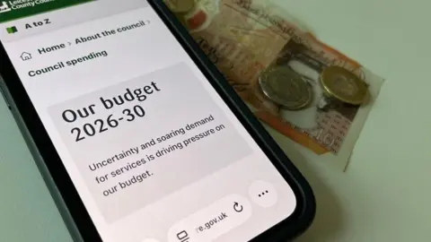 BBC A mobile phone showing Leicestershire County Council's budget page and some money