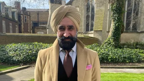 Pharmacy - Max Punni - a man in a tan-coloured turban and coat standing outside looking to the camera