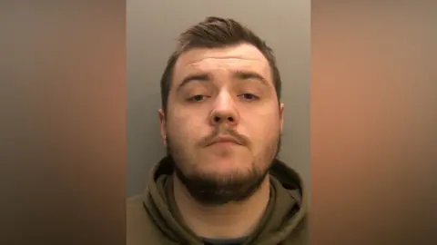 A mugshot of Connor Saggers. He has short brown hair and beard. He has brown eyes and is wearing a olive green hoodie. He is looking at the camera with a serious but somewhat vacant expression.