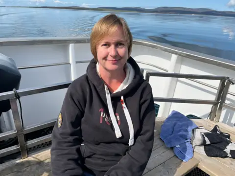 A woman with short blonde hair sitting on a boat with sea and hills in the background.