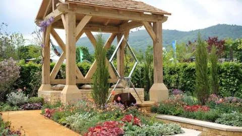 RHS Malvern A garden at last year's event. It includes groups of flowers and small fir trees and a wooden structure. There is also a metal design and a yellow path. 