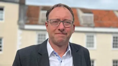 A middle aged man with balding brown hair and glasses looks into the camera, wearing a white shirt and black suit jacket. In the background is a row of houses.