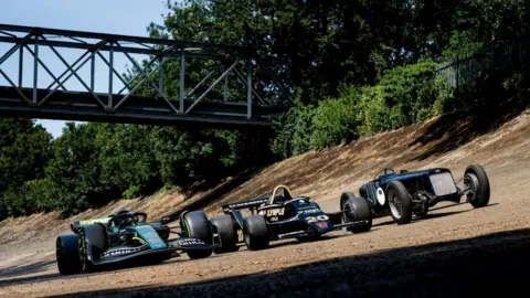Lou Johnson Three racing cars drive alongside one another on a track. Two are modern, one is old fashioned. 