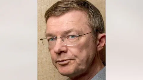 Arne Bye seen on a beige background. He has light hair and blue eyes, and wears glasses.