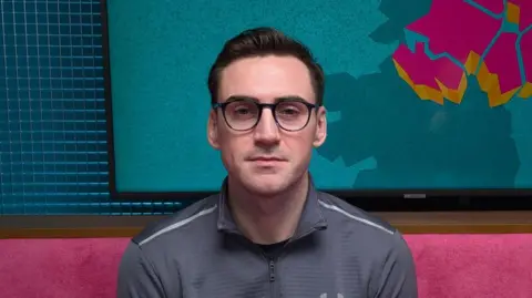 BBC A man is looking straight at the camera. He has short jet black hair, which is slicked back. He has black-rimmed glasses. He is wearing a grey sports top with a zip. He is sitting in a podcast studio with a pink sofa, behind his head is a TV with the logo for The State of Us podcast, which is a teal background with a stylised image of Northern Ireland in pink.