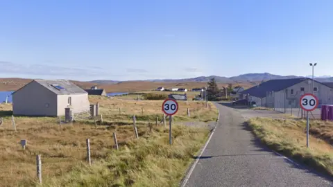 Google A rural scene showing fields with some small buildings and a body of water in the distance. A road connects the buildings, with two 30mph signs visible in the foreground.