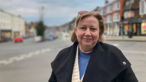 Kym-Marie Cleasby looking into the camera in front of the High Street wearing a black winter coat.