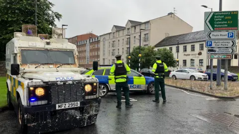 Police vehicles seal off a road in Londonderry. Two officers can be seen beside the vehicles in uniform, including high viz jackets. A number of buildings are in the background