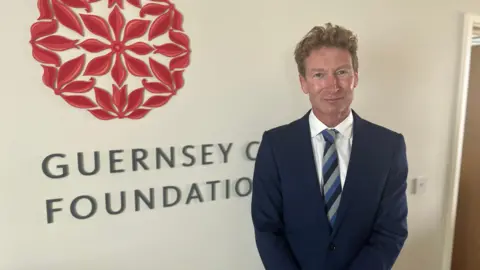 Jim Roberts - A man with wavy ginger hair, wearing a smart blue suit with a white shirt and a blue, black and grey striped tie. He is standing in front of the logo for the Guernsey Community Foundation, with a logo above that is red and looks like a growing flower.